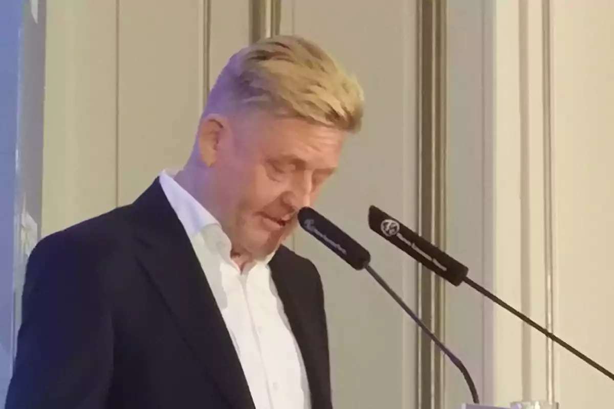 Hombre blanco con traje en una conferencia hablando a dos microfonos