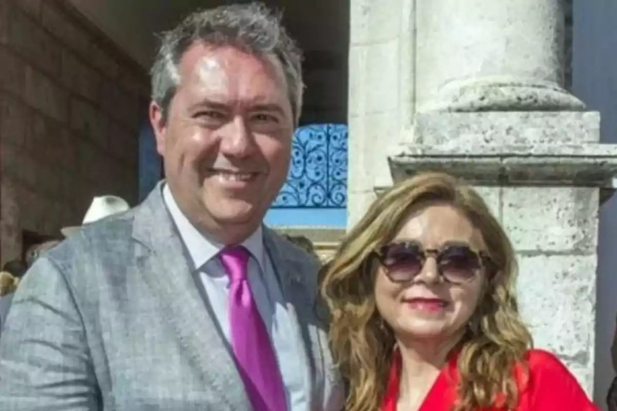 Un hombre con traje gris y corbata rosa sonríe junto a una mujer con gafas de sol y blusa roja frente a una columna de piedra en un día soleado. Un hombre con traje gris y corbata rosa sonríe junto a una mujer con gafas de sol y blusa roja frente a una columna de piedra en un día soleado.