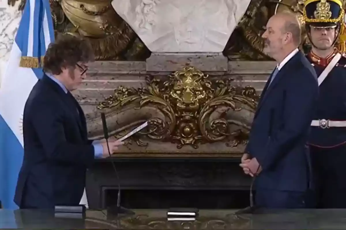 Dos hombres de traje en una ceremonia oficial frente a un busto y una bandera argentina.