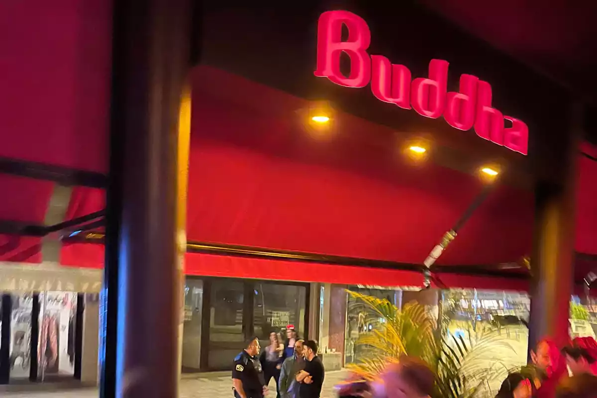 Entrada de un establecimiento con un letrero que dice "Buddha" en letras rojas iluminadas, con varias personas y un oficial de policía en la acera.