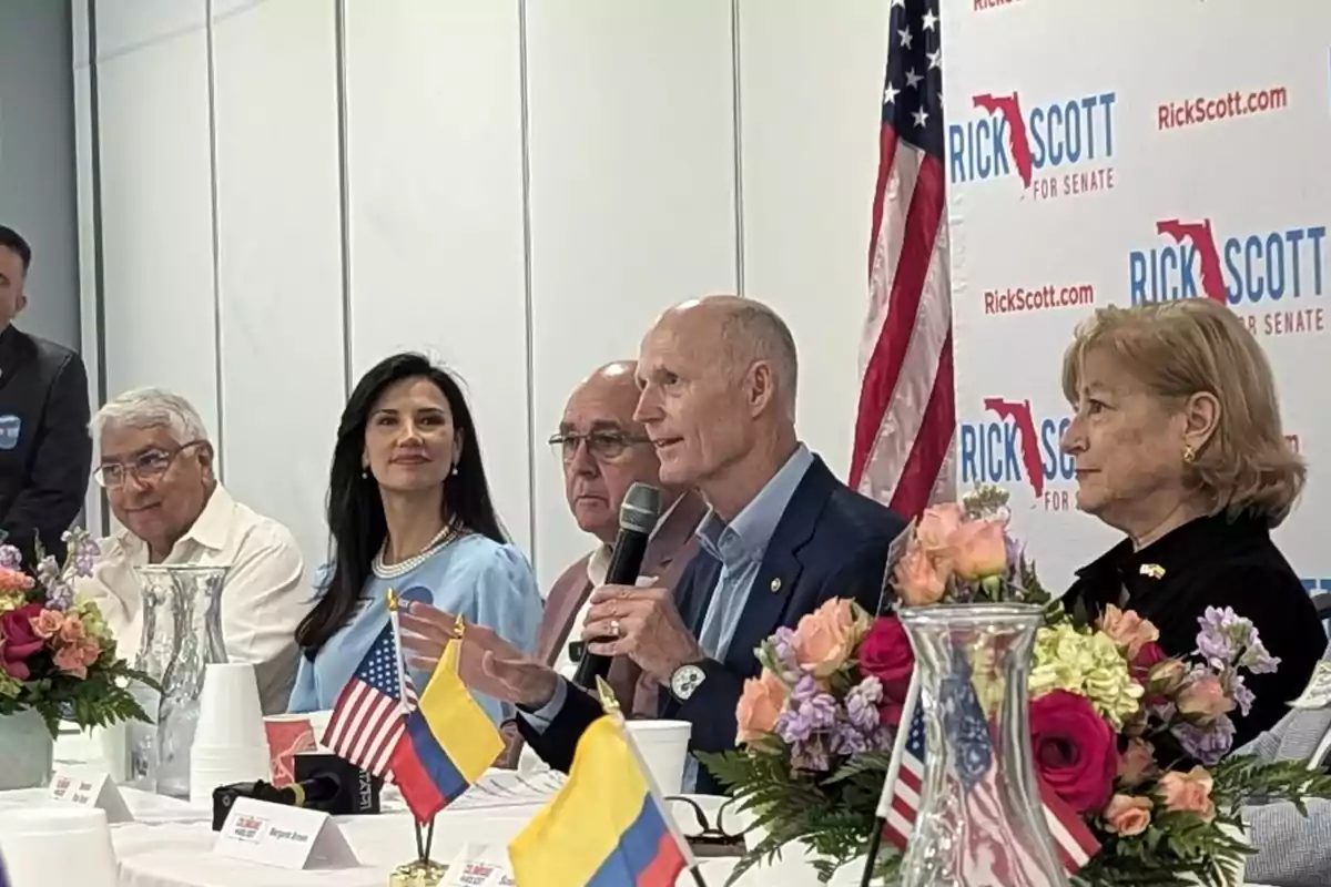 Un grupo de personas está sentado en una mesa decorada con flores y banderas de Estados Unidos y Colombia, mientras una persona habla con un micrófono en la mano; al fondo se ve una bandera estadounidense y un cartel de campaña política.