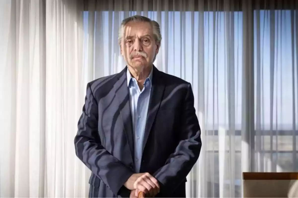 Hombre mayor con traje oscuro y camisa azul claro, de pie frente a una cortina blanca con luz natural.