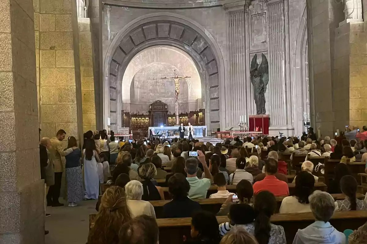 Una multitud de personas sentadas en bancos dentro de una iglesia, observando una ceremonia frente al altar con una gran cruz y una estatua de ángel en el fondo.