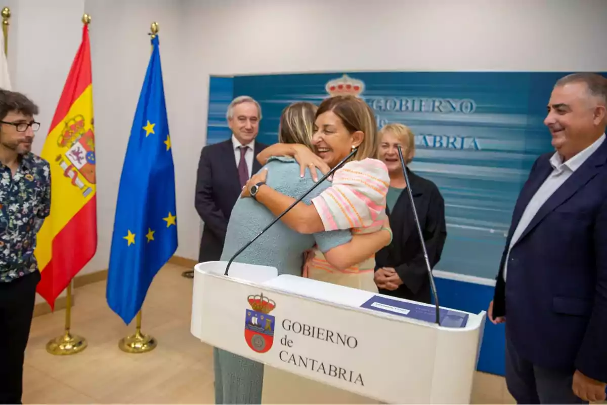 Personas abrazándose en un evento oficial del Gobierno de Cantabria, con banderas de España y la Unión Europea en el fondo.