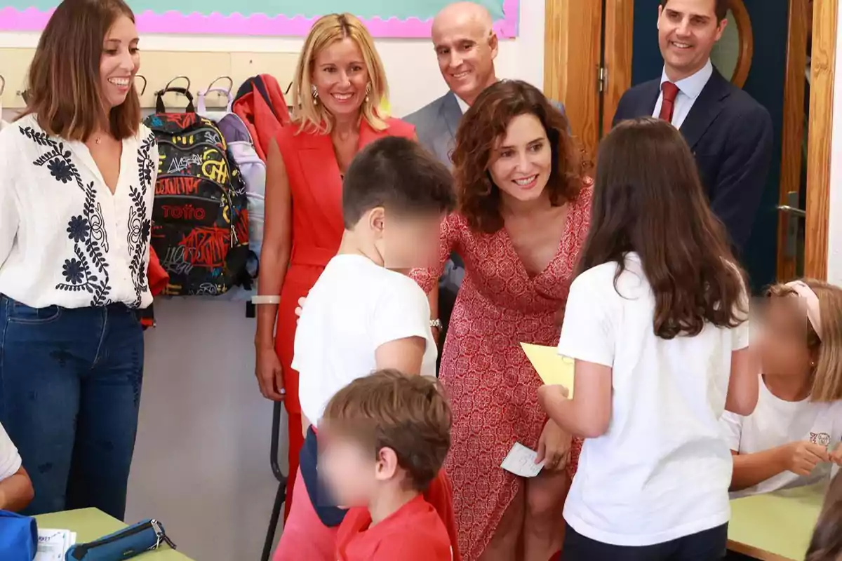 Un grupo de adultos y niños interactuando en un aula escolar, con mochilas colgadas en el fondo y una mujer de vestido rojo inclinada hacia los niños.