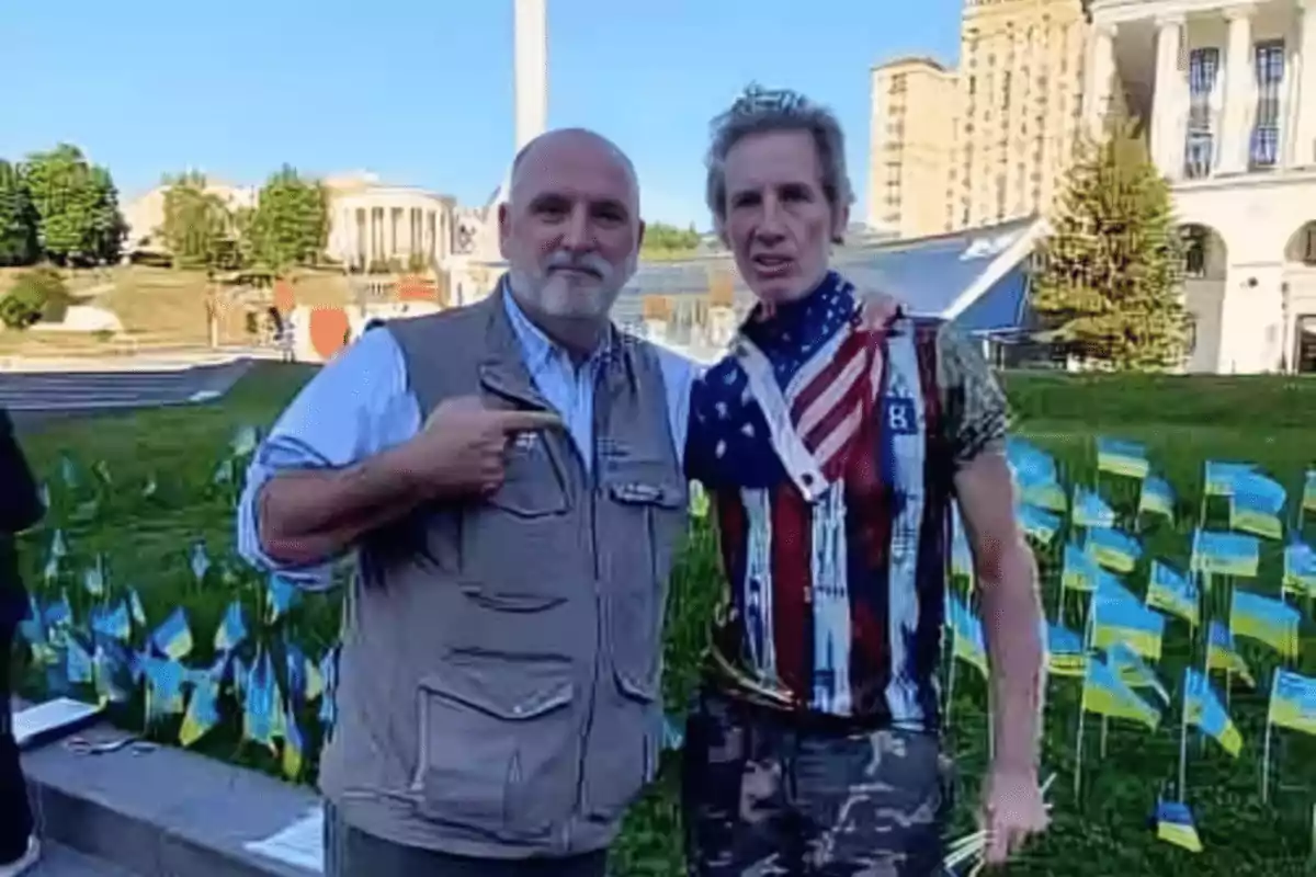 Two people pose together outdoors, one of them wearing a T-jersey with the American flag and the other pointing toward him, in the background there is a building and several small Ukrainian flags on the grass. Two people pose together outdoors, one of them wearing a T-jersey with the American flag and the other pointing toward him, in the background there is a building and several small Ukrainian flags on the grass.