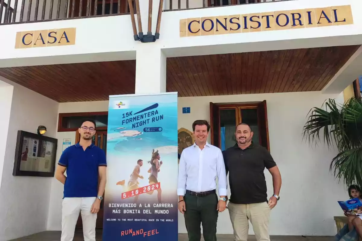 Tres hombres posan frente a un edificio con un cartel que anuncia la "15K Formentera Night Run" el 5 de octubre de 2024.