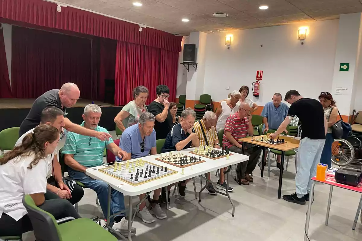 Un grupo de personas de diferentes edades juega al ajedrez en una sala con cortinas rojas y sillas verdes, mientras otras observan y participan en la actividad.