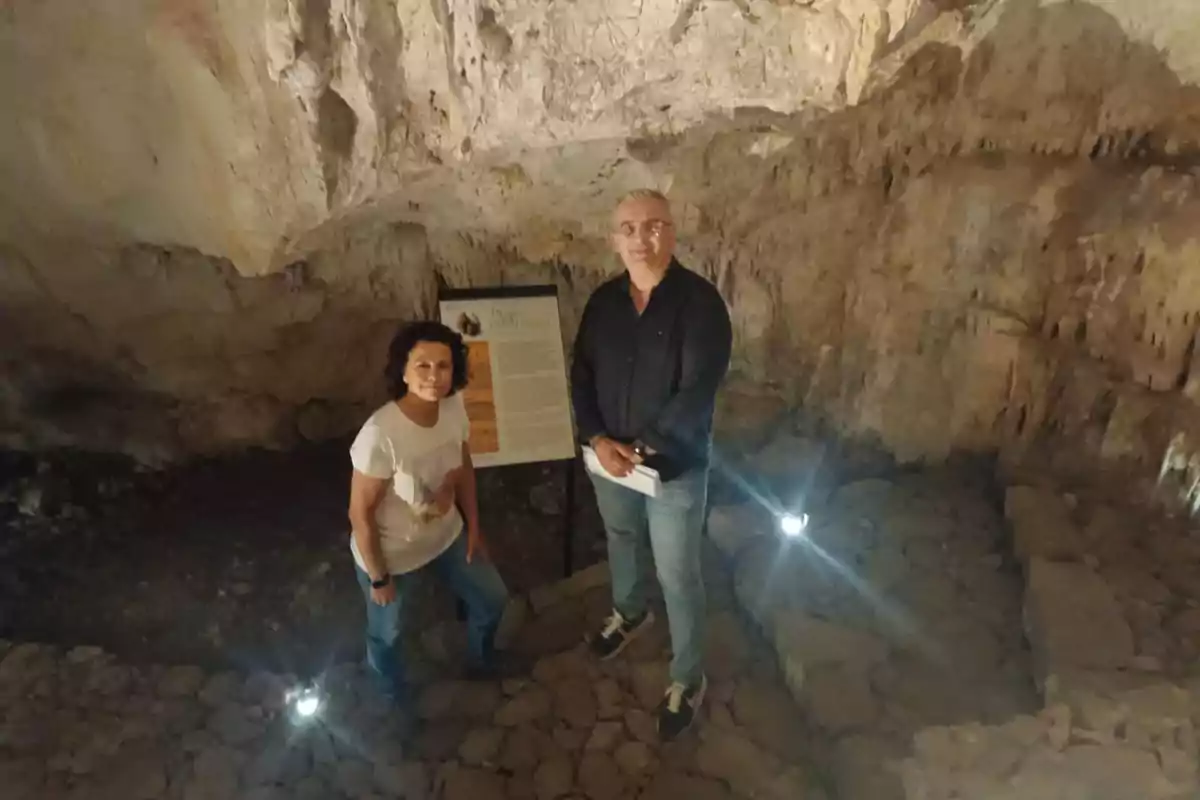 Dos personas posan dentro de una cueva iluminada, junto a un cartel informativo.