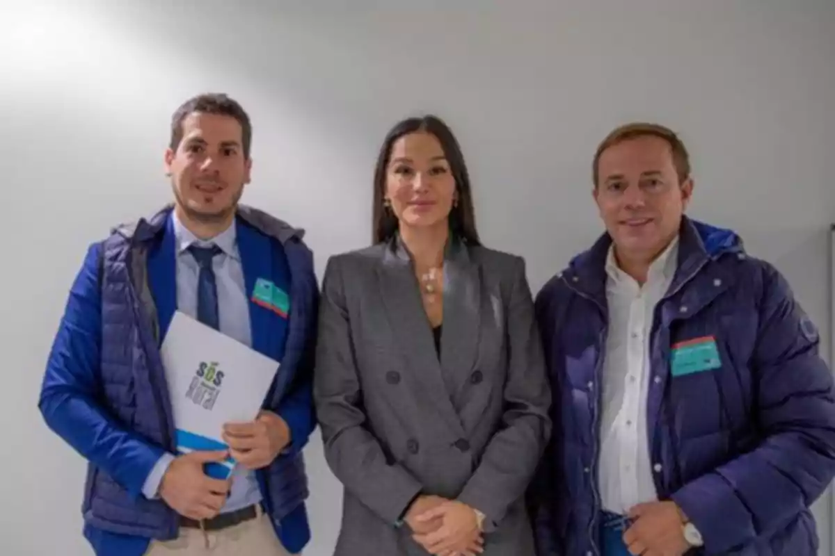 Tres personas posan juntas frente a una pared blanca, dos hombres con chaquetas azules y una mujer con un traje gris.