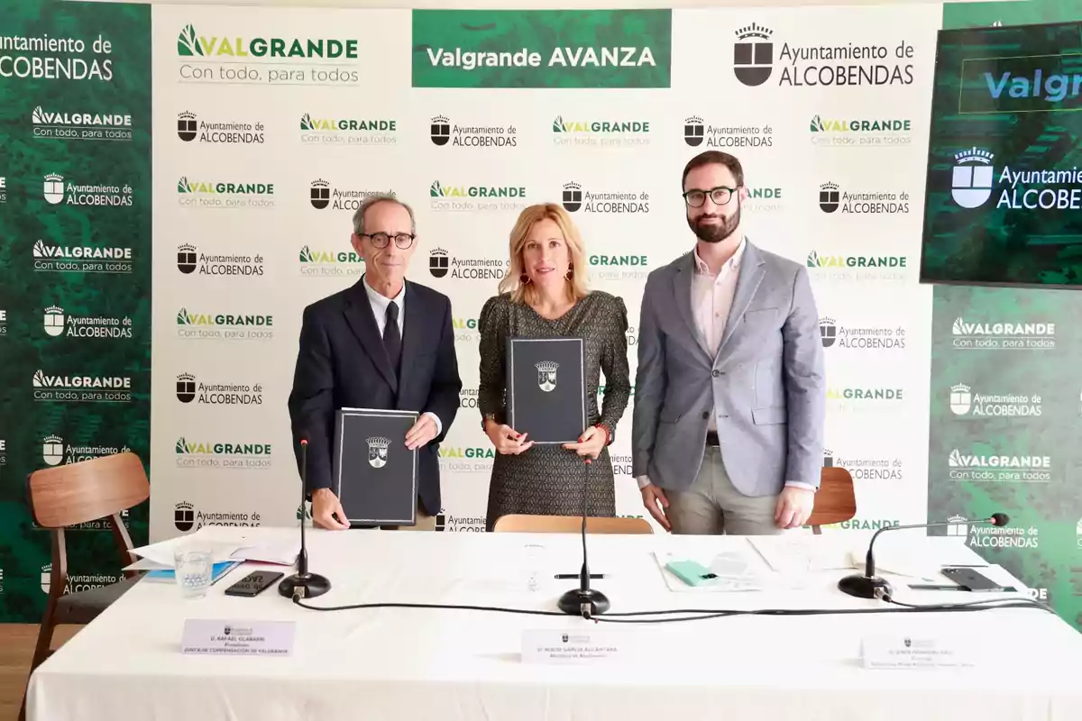 Tres personas posan con carpetas en un evento del Ayuntamiento de Alcobendas con el logo de Valgrande en el fondo.