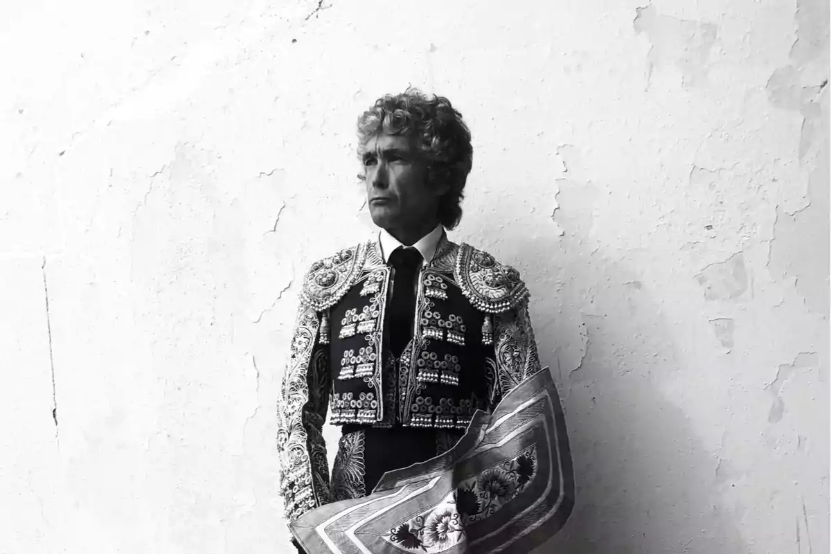 Un torero con traje tradicional y capote en blanco y negro frente a una pared texturizada.