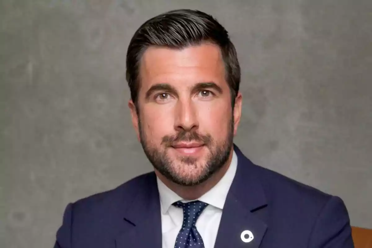 Un hombre con barba y cabello oscuro lleva un traje azul y corbata, posando frente a un fondo gris.