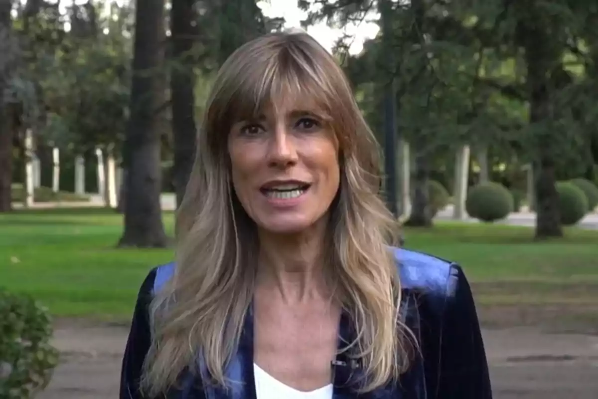 Una mujer de cabello rubio y largo está de pie en un parque con árboles y arbustos al fondo.