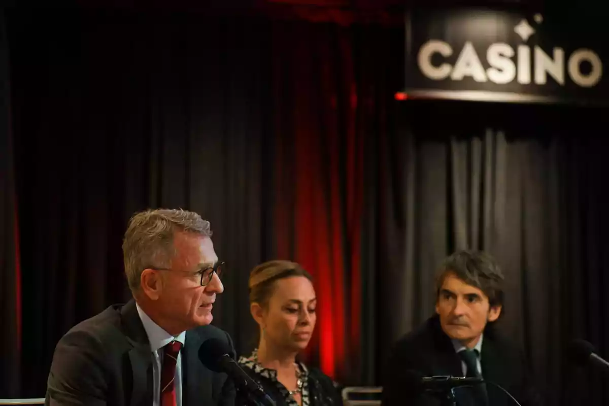 Tres personas sentadas en una mesa con micrófonos en un evento en un casino.