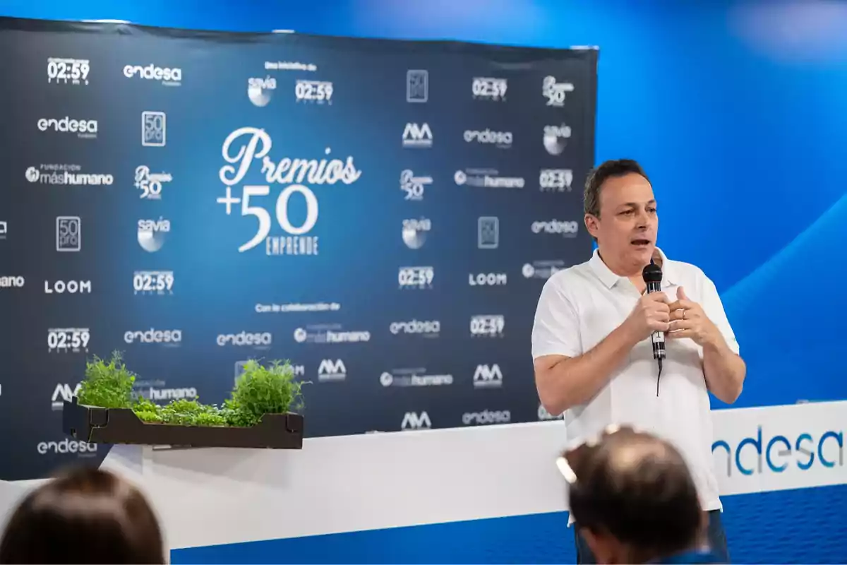 Un hombre hablando con un micrófono en un evento de los Premios +50 Emprende, con un fondo azul que muestra varios logotipos de patrocinadores.