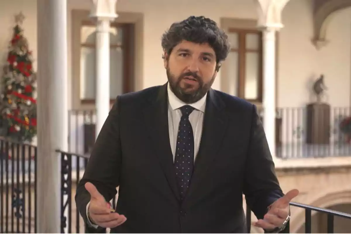 Hombre con traje oscuro y corbata de pie en un balcón decorado con un árbol de Navidad al fondo.