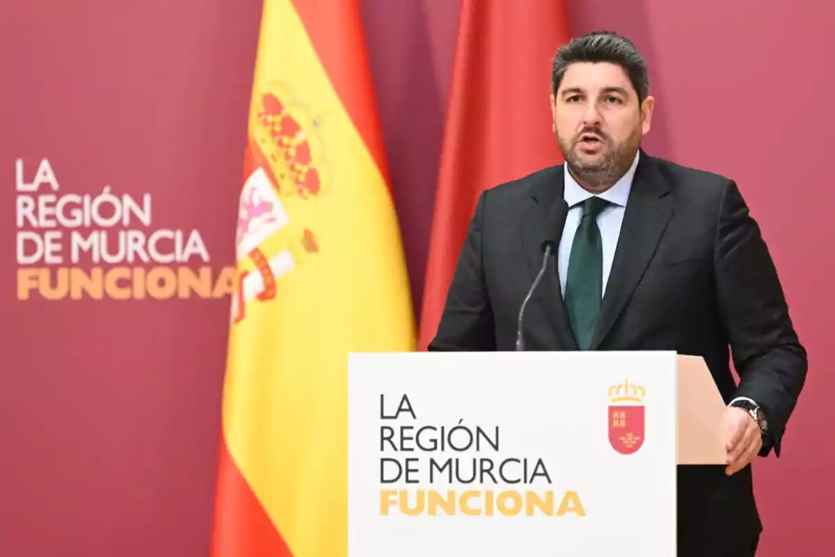 Hombre de traje hablando en un podio con el texto "La Región de Murcia Funciona" y una bandera de España al fondo.