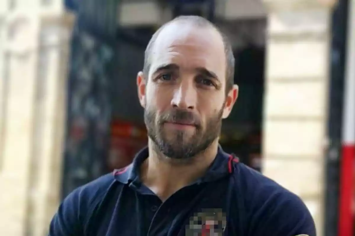 Un hombre con barba y cabello corto viste una camiseta oscura y está de pie frente a un fondo urbano desenfocado.