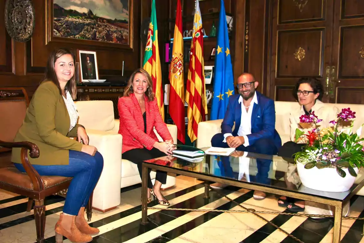 Four people seated in a meeting room with flags and a painting on the wall, around a glass table with a floral arrangement.