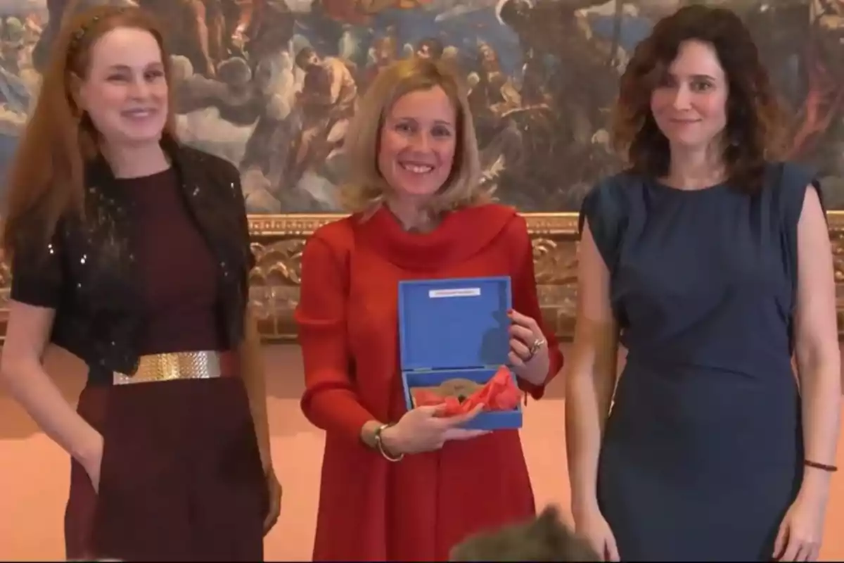 Tres mujeres posan juntas, una de ellas sostiene un estuche azul con un objeto en su interior, y están frente a una pintura enmarcada.