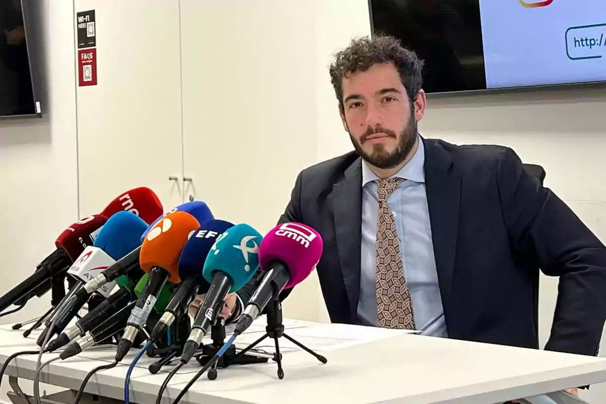 A man in a suit and tie is sitting in front of a table full of microphones from different media outlets during a press conference.