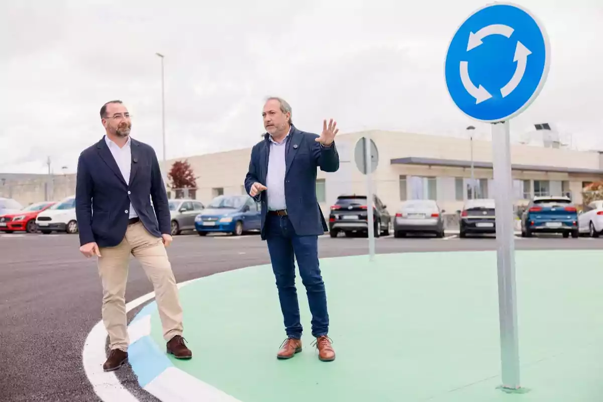 Dos hombres de pie en una rotonda junto a una señal de tráfico azul con flechas circulares, con varios coches estacionados y un edificio al fondo.