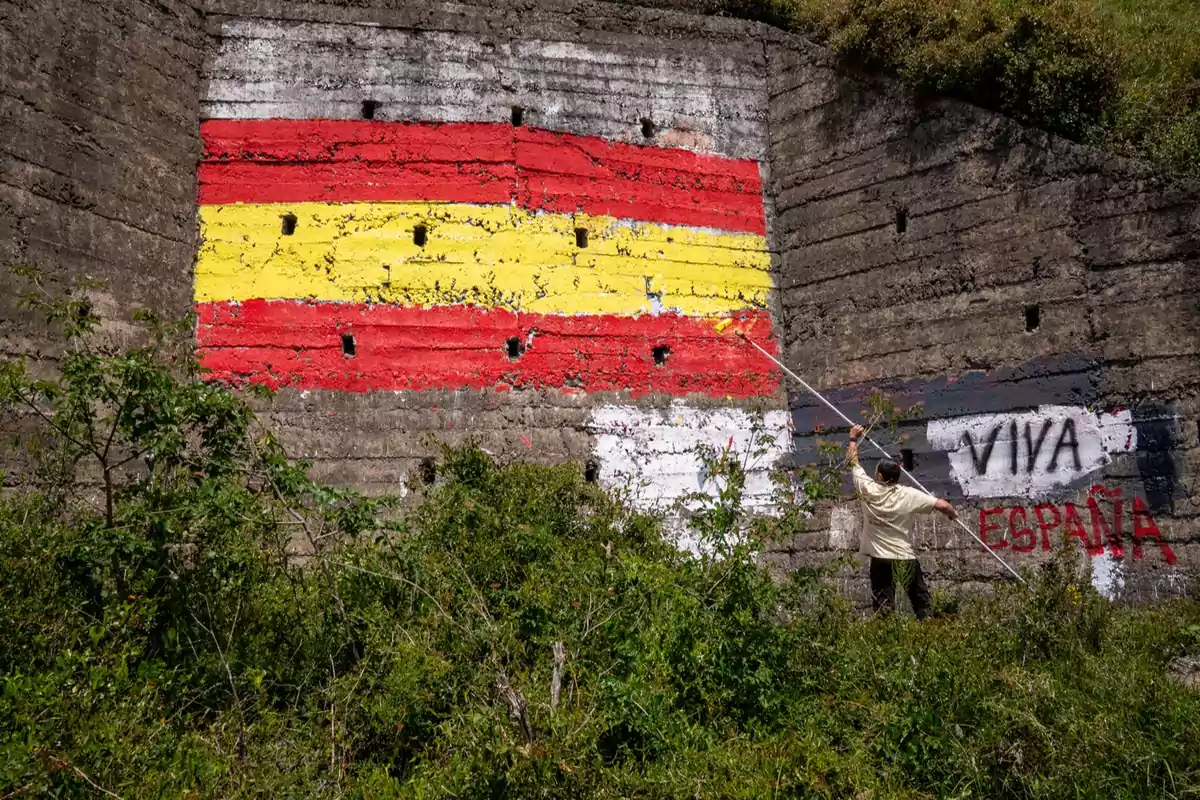 Persona pintando una bandera de España en una pared de cemento junto a un grafiti que dice Viva España rodeado de vegetación