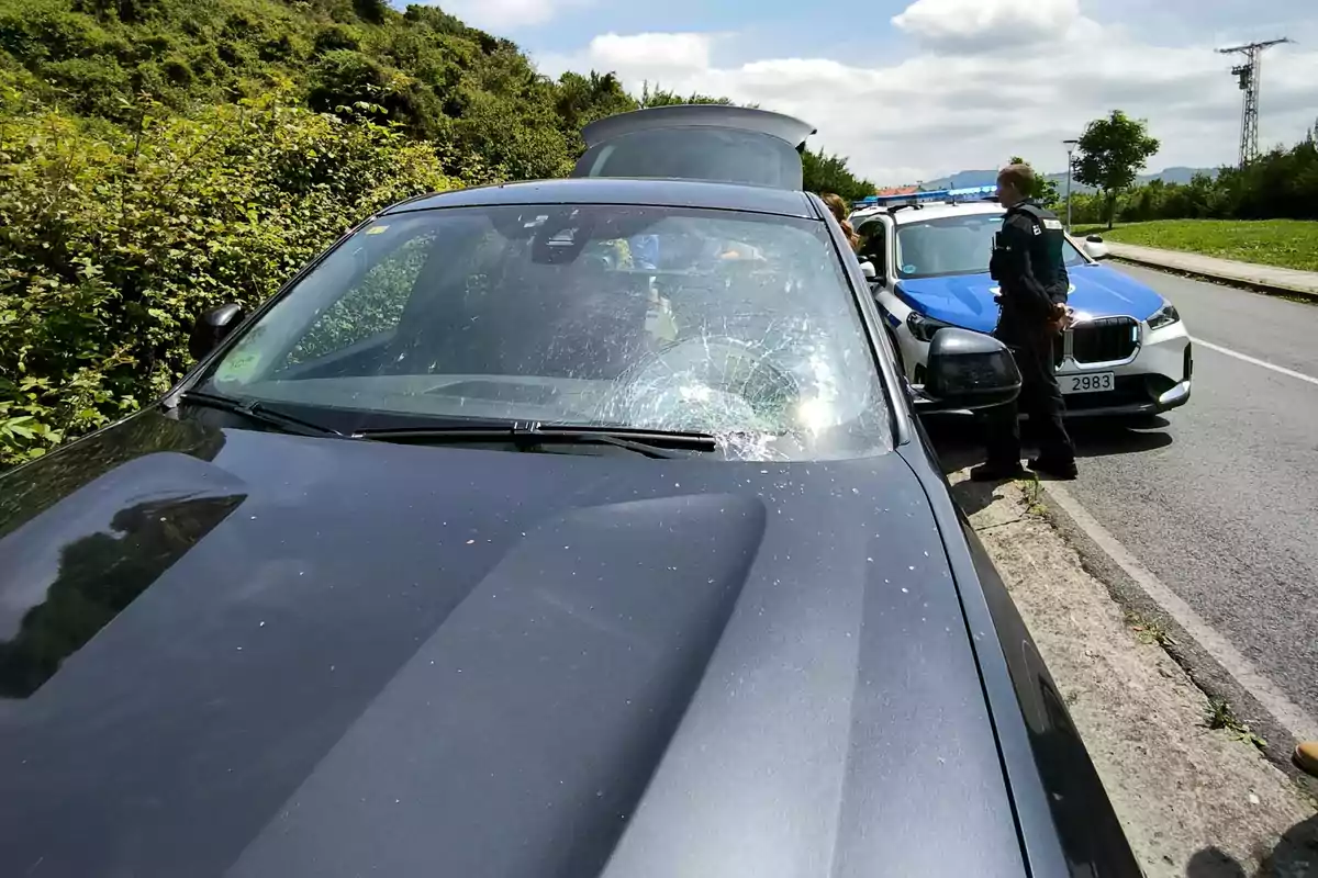 Un coche negro con el parabrisas roto está detenido al lado de una carretera junto a un coche de policía y un agente de pie Un coche negro con el parabrisas roto está detenido al lado de una carretera junto a un coche de policía y un agente de pie