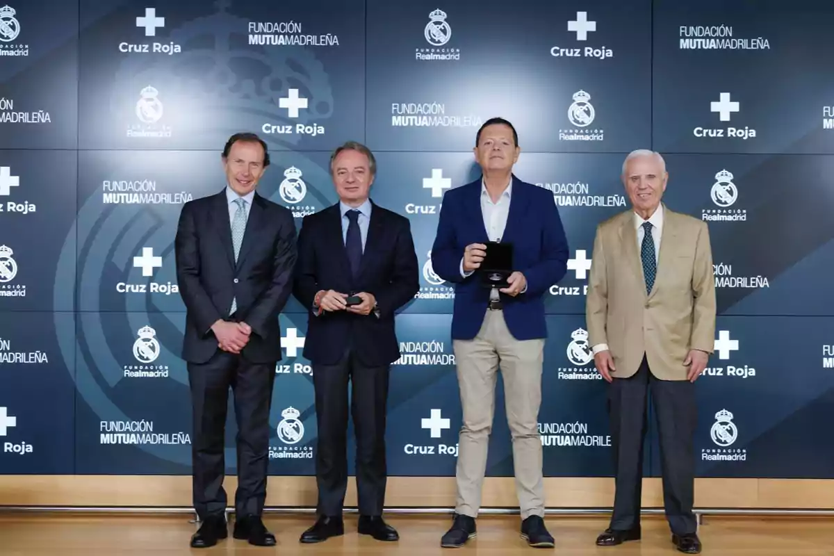 Cuatro hombres posan de pie en un evento formal frente a un fondo con logotipos de la Fundación Real Madrid, Cruz Roja y Fundación Mutua Madrileña