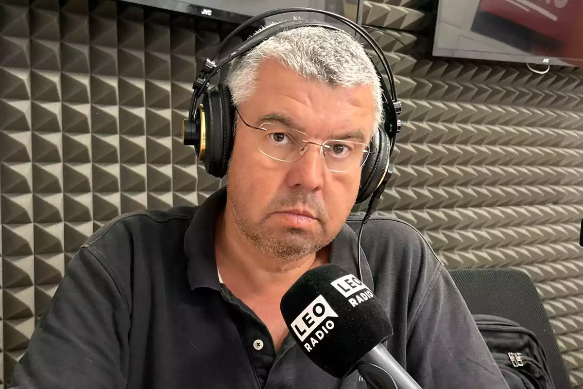 Hombre de cabello canoso y gafas usando audífonos frente a un micrófono en una cabina de radio