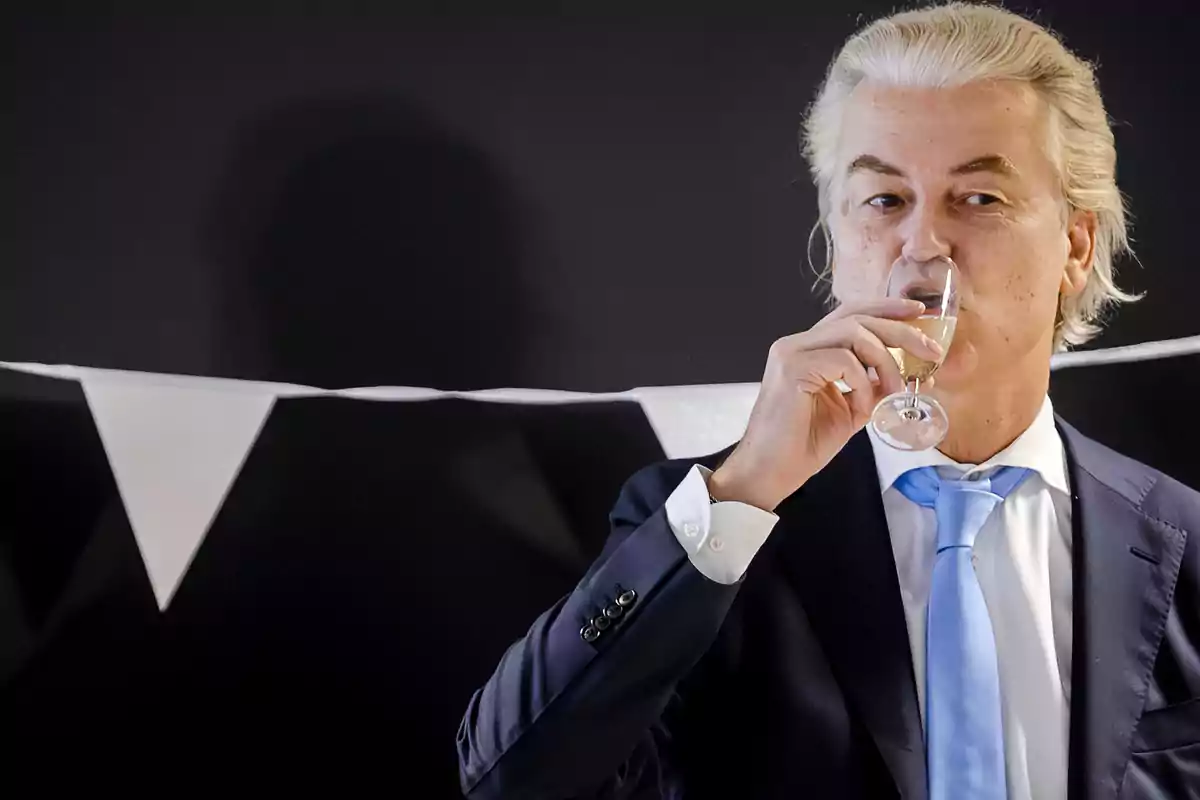 Hombre de cabello canoso con traje azul y corbata celeste bebiendo de una copa de vino espumoso frente a una guirnalda de banderines blancos y negros Hombre de cabello canoso con traje azul y corbata celeste bebiendo de una copa de vino espumoso frente a una guirnalda de banderines blancos y negros