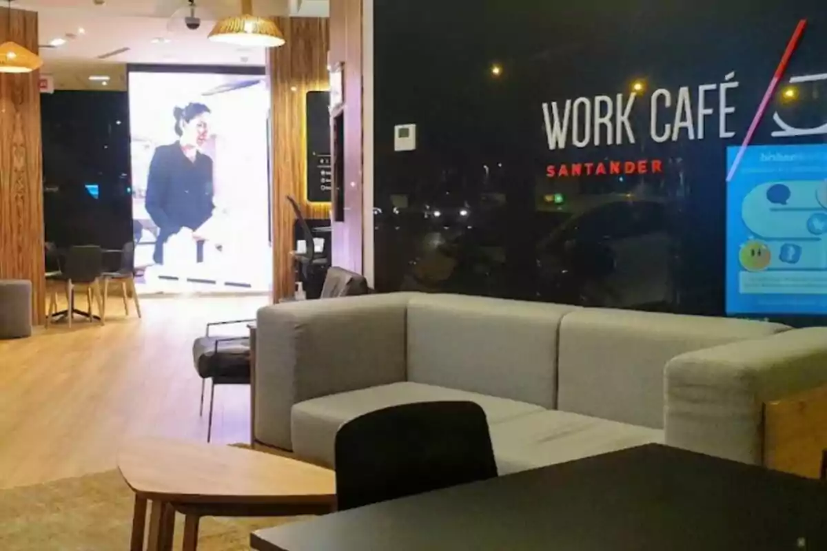 La imagen muestra el interior de un Work Café Santander, con un sofá gris, una mesa de madera y sillas en un ambiente moderno y bien iluminado. La imagen muestra el interior de un Work Café Santander, con un sofá gris, una mesa de madera y sillas en un ambiente moderno y bien iluminado.