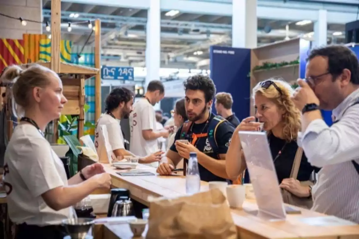 Personas interactuando en un stand de una feria o evento, algunas probando bebidas y otras atendiendo detrás del mostrador.