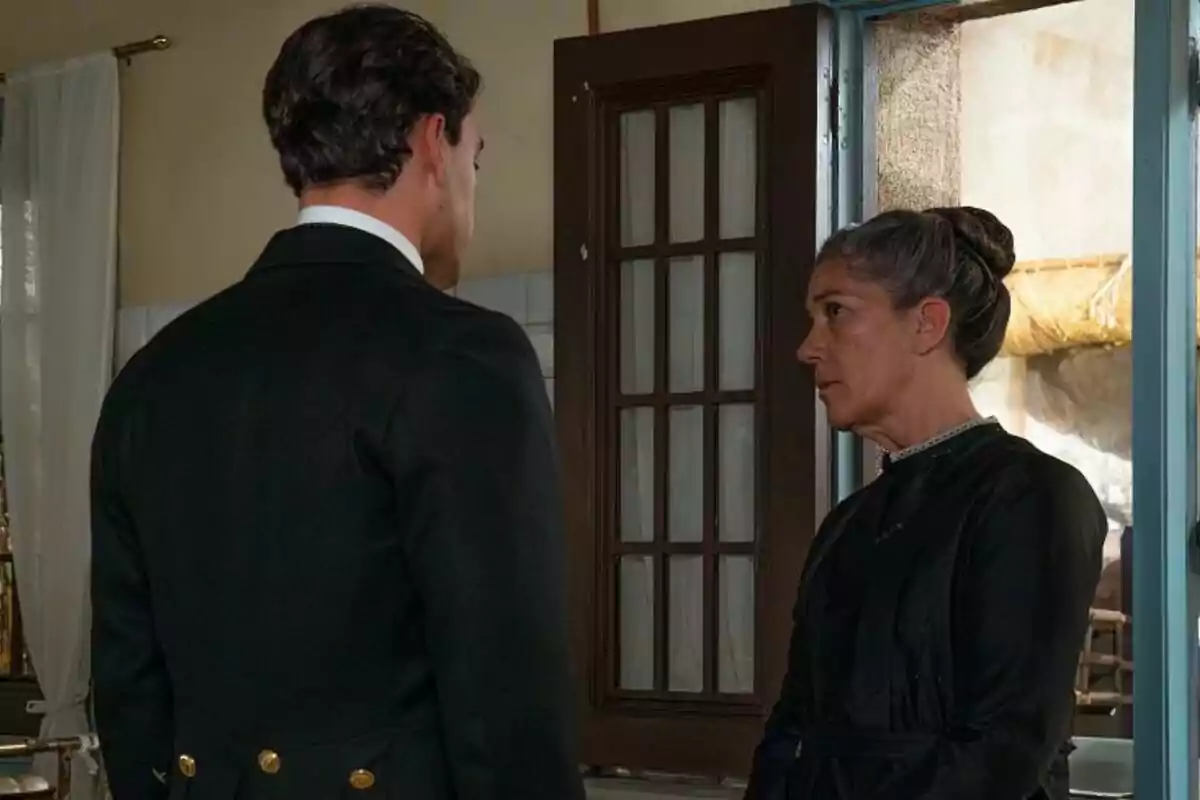 Xavi Lock y Marga Martínez están de pie frente a una puerta, mirándose con expresiones serias en una habitación iluminada en La Promesa. Xavi Lock y Marga Martínez están de pie frente a una puerta, mirándose con expresiones serias en una habitación iluminada en La Promesa.