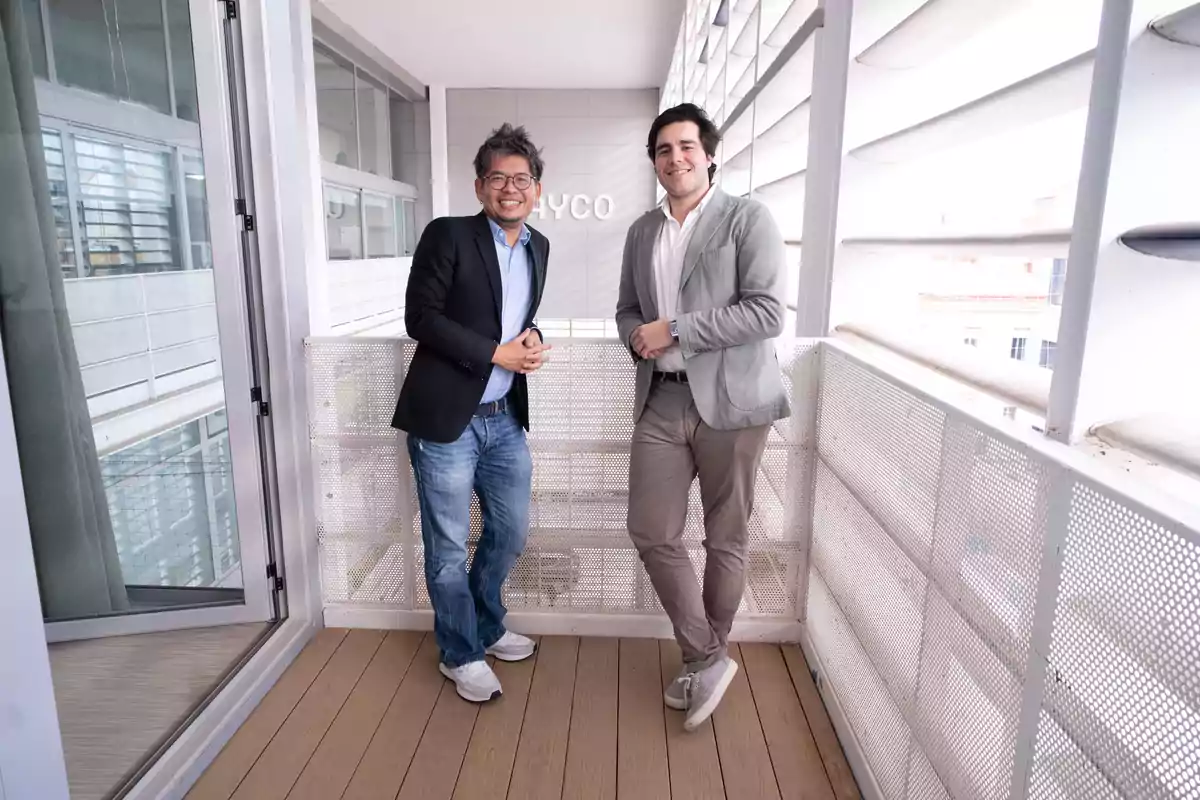 Dos hombres de pie en un balcón moderno con barandilla de metal y fondo de edificio.