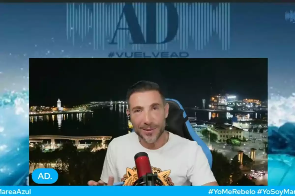 Antonio David Flores con camiseta clara hablando frente a un micrófono rojo con fondo nocturno de una ciudad iluminada y el mar Antonio David Flores con camiseta clara hablando frente a un micrófono rojo con fondo nocturno de una ciudad iluminada y el mar