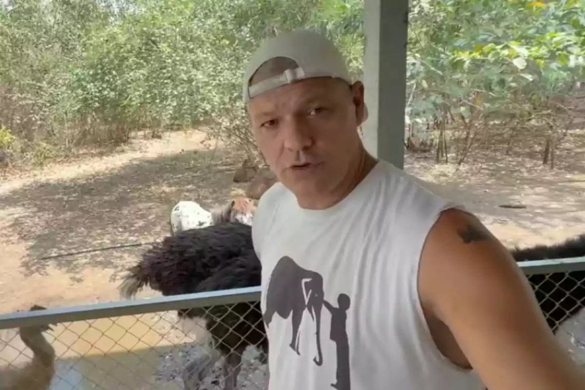 Frank Cuesta con gorra y camiseta sin mangas junto a un avestruz detrás de una cerca en un entorno natural. Frank Cuesta con gorra y camiseta sin mangas junto a un avestruz detrás de una cerca en un entorno natural.