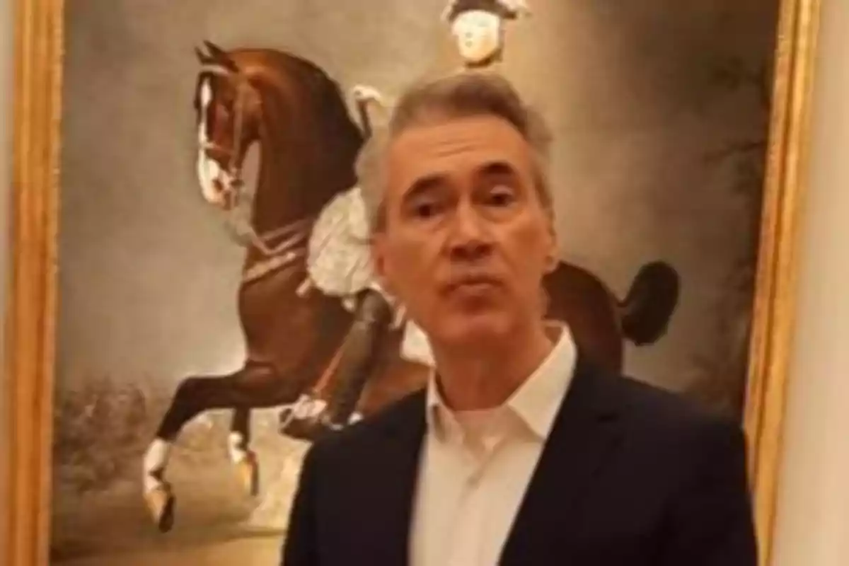 Gray-haired man wearing a dark jacket and white jersey posing in front of a painting of a rider on a horse Gray-haired man wearing a dark jacket and white jersey posing in front of a painting of a rider on a horse