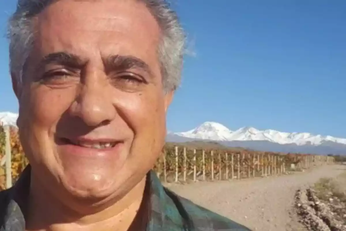 Hombre sonriendo con montañas nevadas y viñedos al fondo en un día soleado.