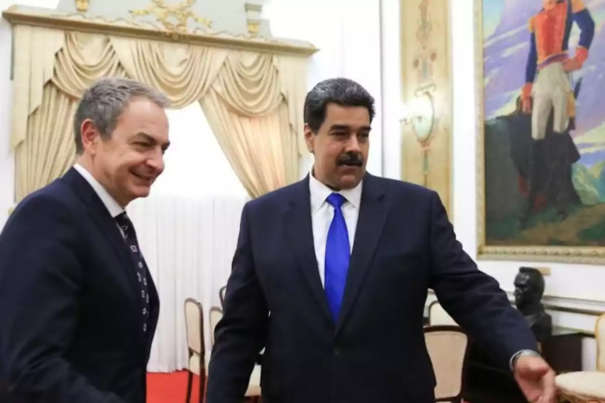 Dos hombres de traje conversan en una sala decorada con cortinas elegantes y un gran cuadro en la pared.