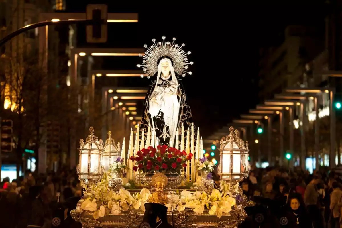 Procesión nocturna con una imagen religiosa adornada con flores y velas rodeada de una multitud en una calle iluminada.