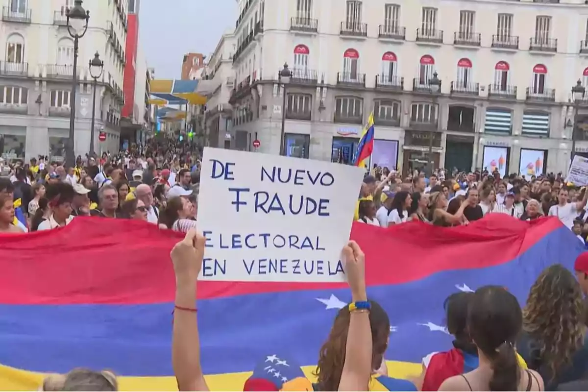 Una multitud de personas se manifiesta en una calle, sosteniendo una gran bandera de Venezuela y un cartel que dice "De nuevo fraude electoral en Venezuela".